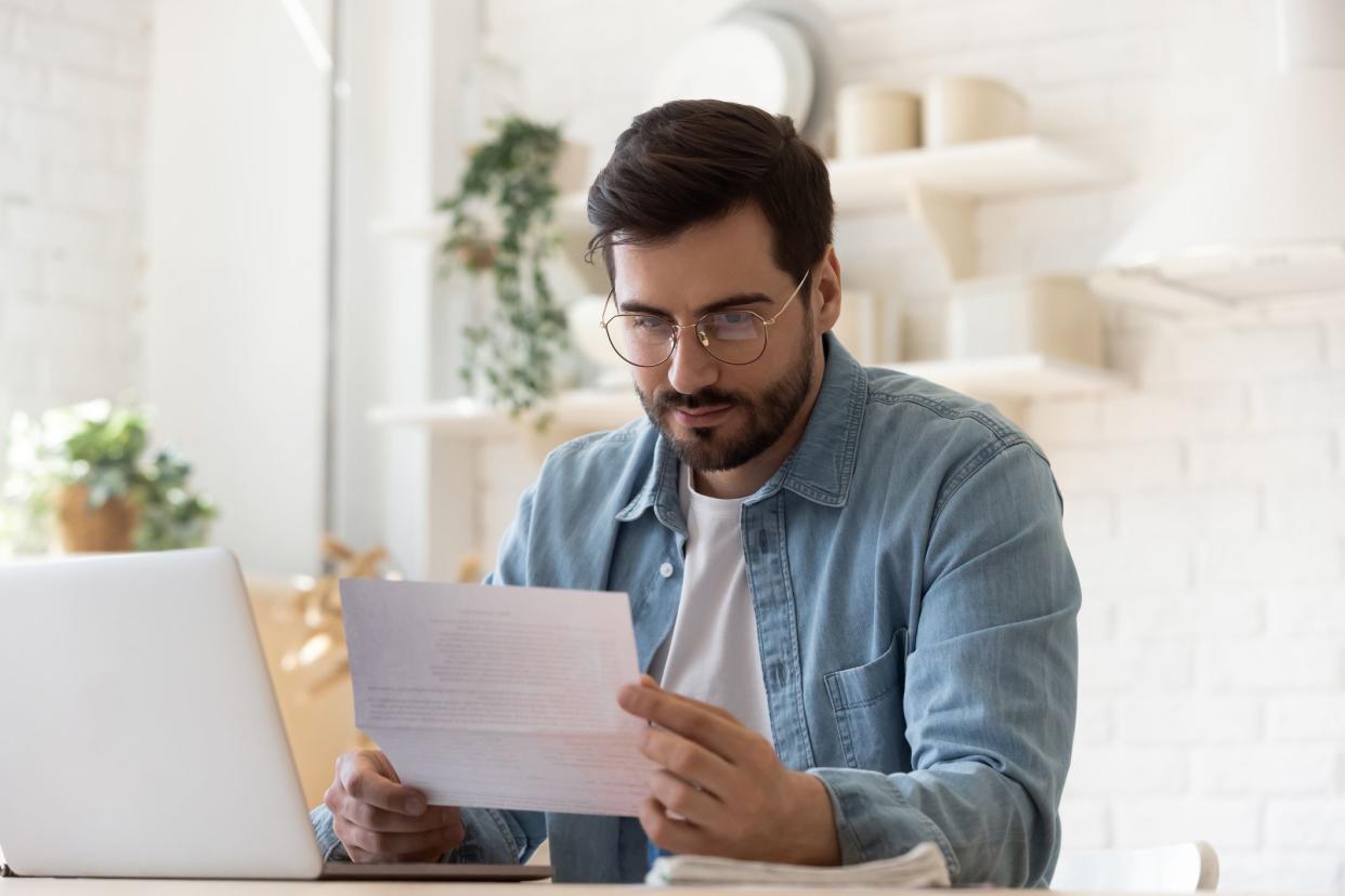 Pensive millennial man in glasses sit at table work on laptop read letter correspondence thinking, thoughtful young male distracted from computer consider paperwork or post notification at home