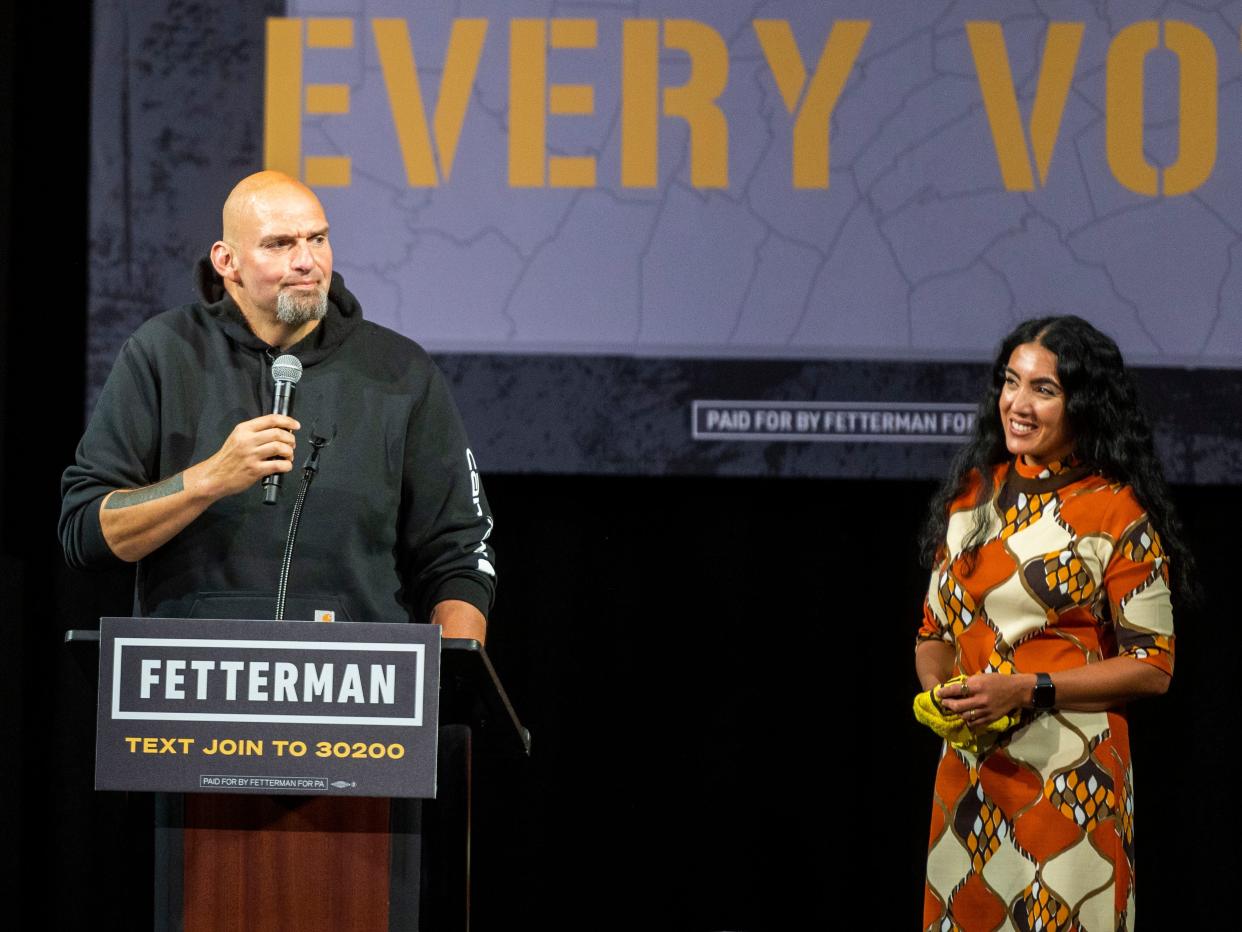 John Fetterman speaks onstage as his wife, Gisele Barreto Fetterman, looks on.