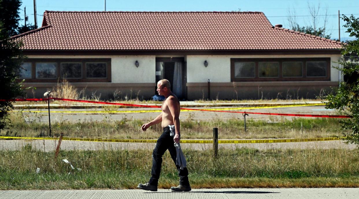 A shirtless man walks in front of a home that appears abandoned. Behind the man and in front of the house are multiple lines of red and yellow police tape warning people not to enter the crime scene. 