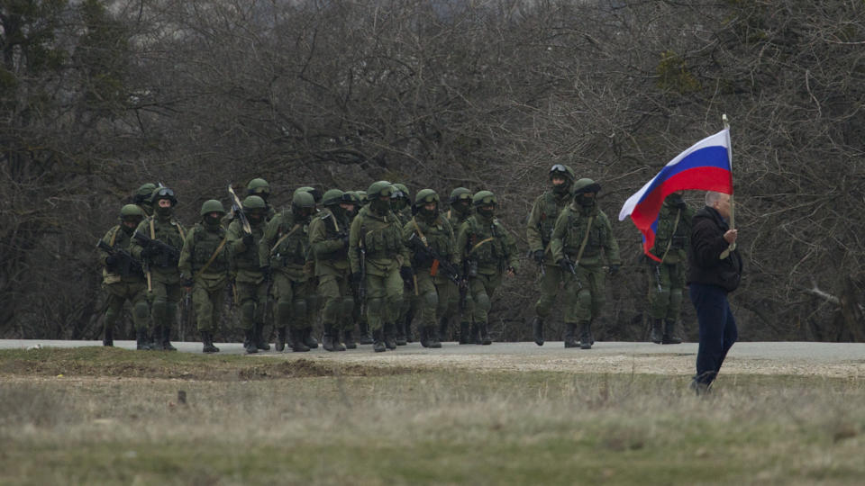 Russian soldiers walk as a local resident waves with Russian flag outside of Ukrainian military base in the village of Perevalne, outside of Simferopol, Ukraine, on Sunday, March 2, 2014. Hundreds of armed men in trucks and armored vehicles surrounded the Ukrainian military base Sunday in Crimea, blocking its soldiers from leaving. (AP Photo/Ivan Sekretarev)