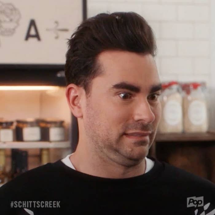 Dan Levy, with a surprised expression, standing in front of a shelf in a scene from Schitt's Creek