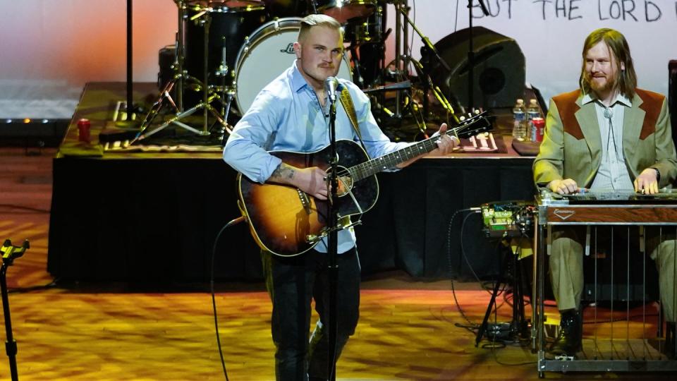 zach bryan playing his guitar while singing as a band plays in the background