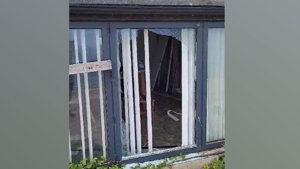 A close-up of black framed three-pane window with the middle pane smashed, showing through into a room beyond