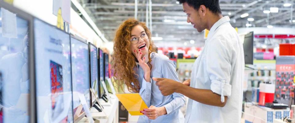 African guy with red-haired girl standing in front of the stand in the electronics store choose a plasma TV looking at the price tags