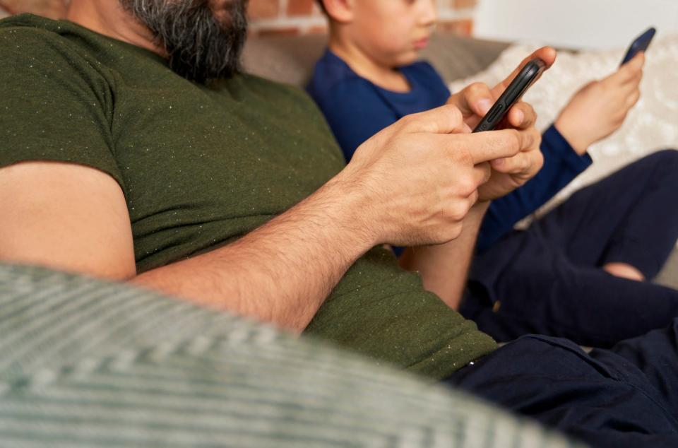 A man with a beard and a boy sit on a couch, both focused on their smartphones. The man wears a short-sleeved shirt, and the boy wears long sleeves