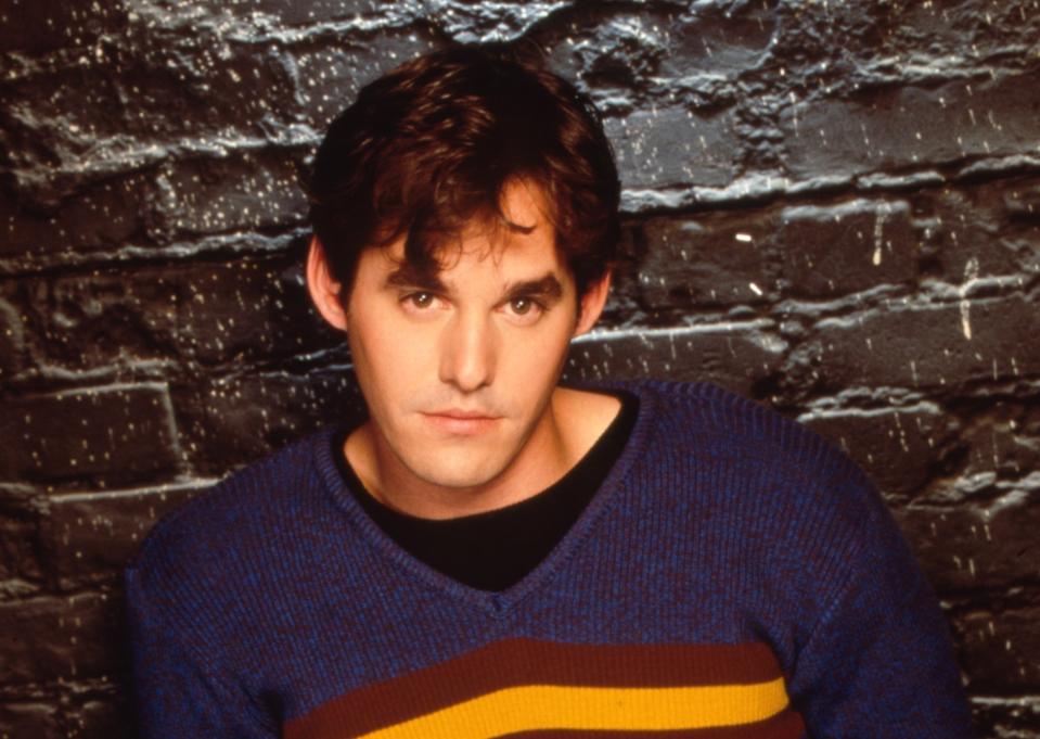 Man in a blue sweater with a yellow and red stripe, leaning against a brick wall