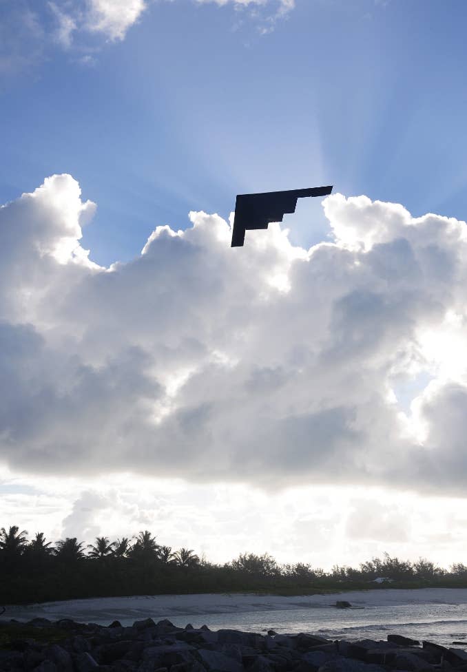 A stealth bomber flies overhead against a backdrop of clouds and sunrays, with a distant treeline and body of water visible below