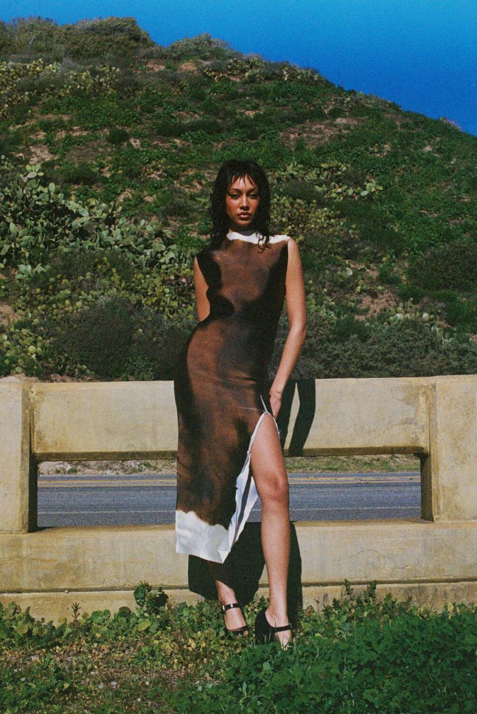 Model wears a black dress in the sunshine, a green hill behind her