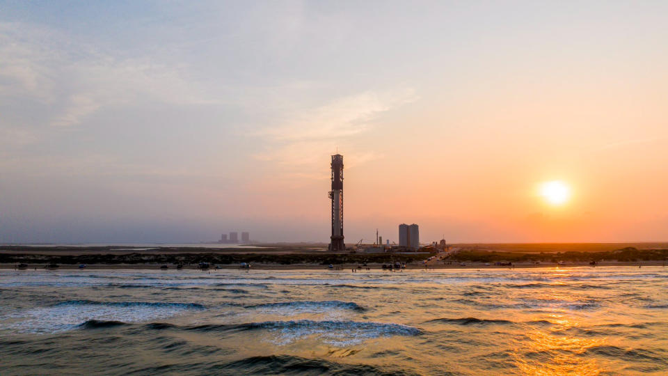 view of a giant rocket on the launch pad with the ocean in the foreground and the setting sun in the background