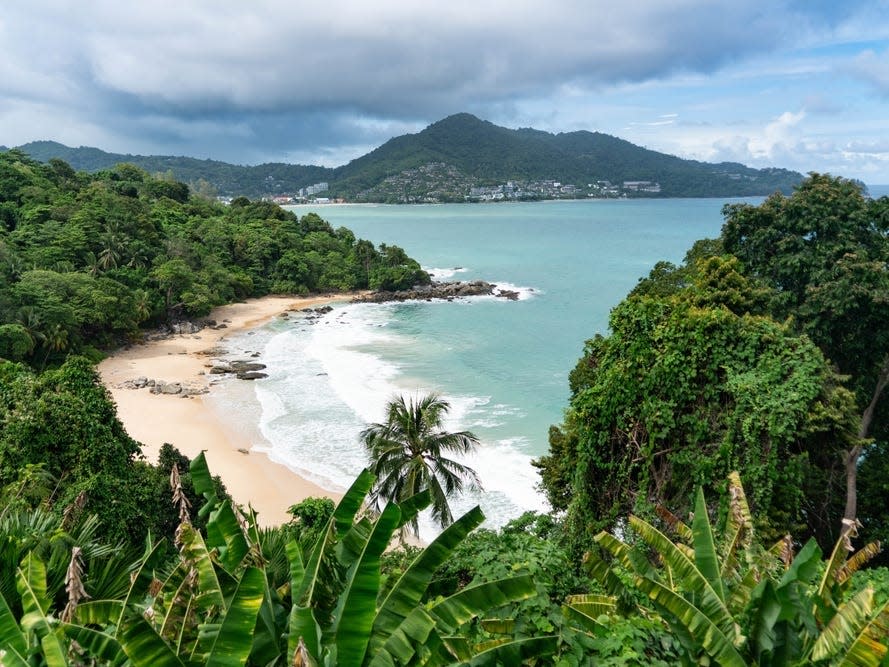 Ein Strand mit üppigem Grün, türkisfarbenem Wasser und Bergen in der Ferne.