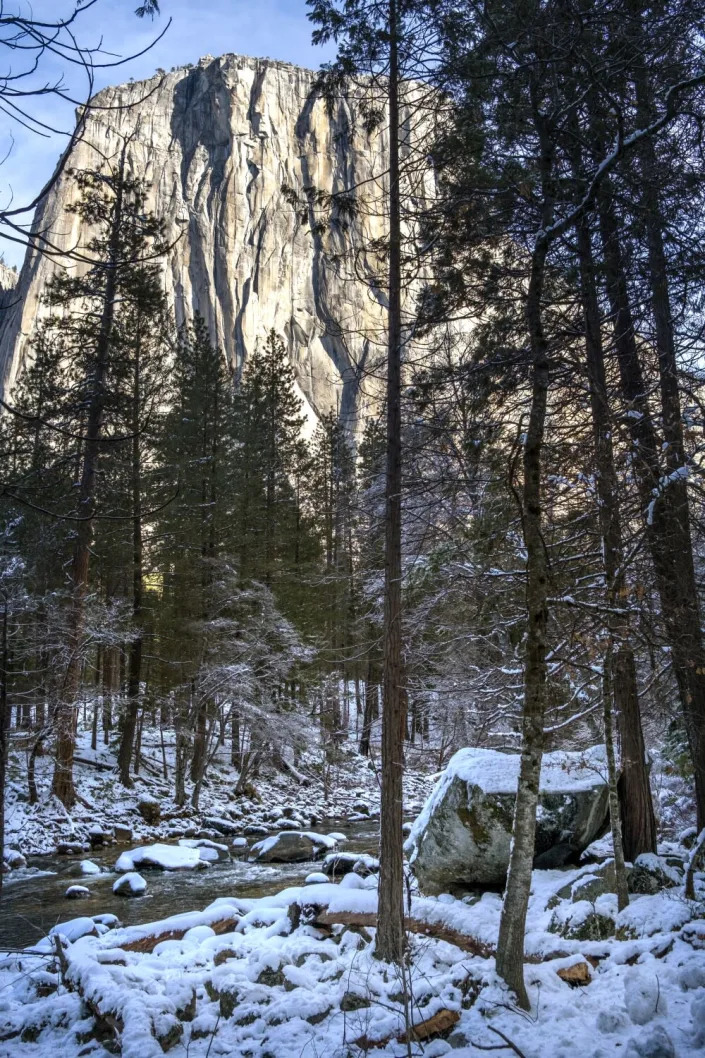 A large granite prominence towers behind sparse conifers lining a stream with snowy banks.