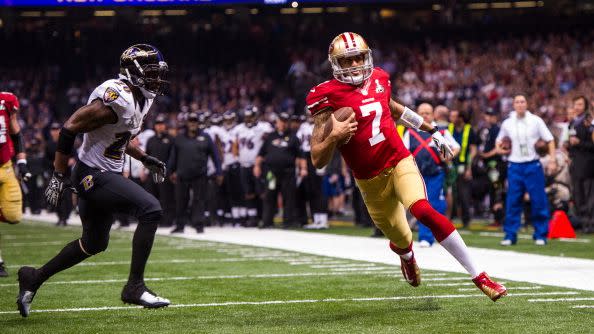 colin kaepernick runs on a football field just outside a red endzone while holding a football and wearing a san francisco 49ers uniform, a player for the baltimore ravens runs toward him from the left, ravens players and staff stand in the background and fans watch from stands