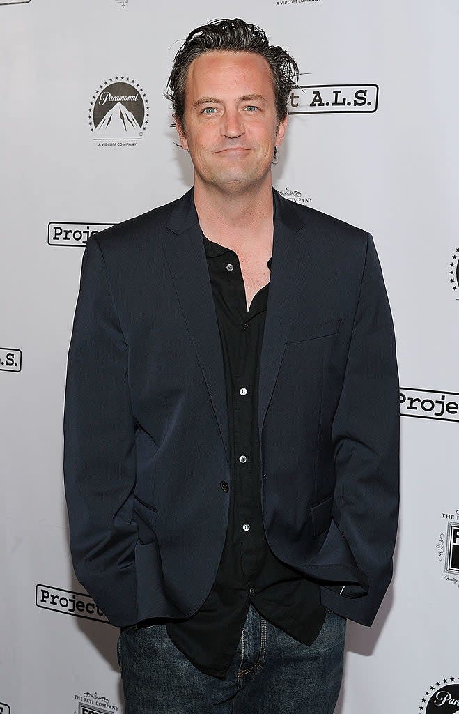 Matthew Perry standing, wearing a suit jacket over an unbuttoned shirt