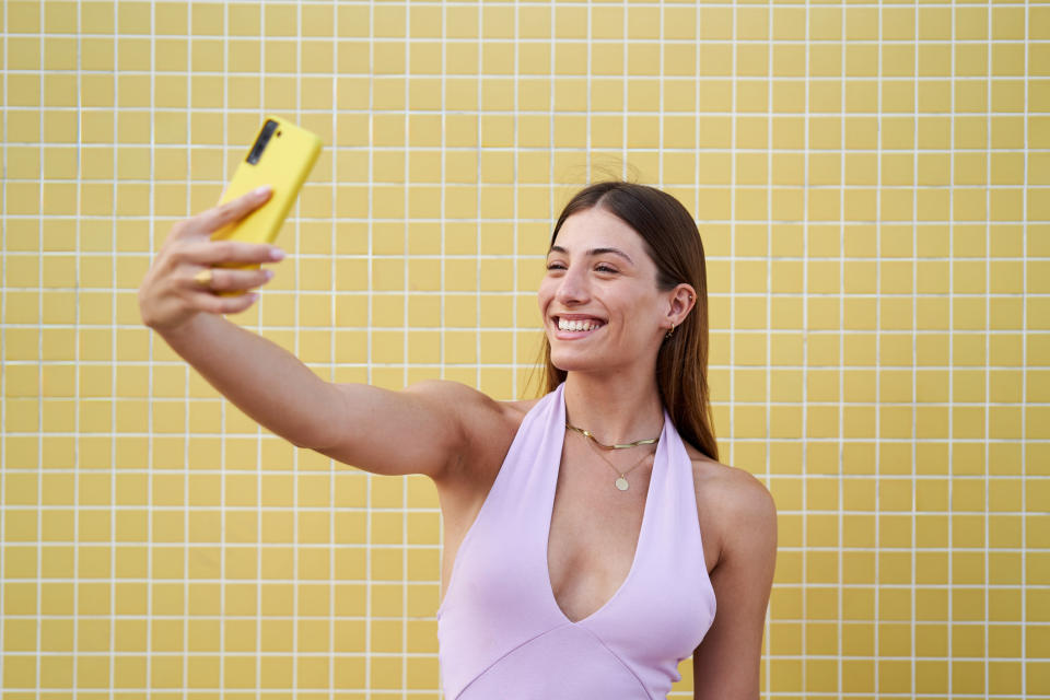 A woman in a sleeveless top smiles and takes a selfie with her yellow phone against a tiled background