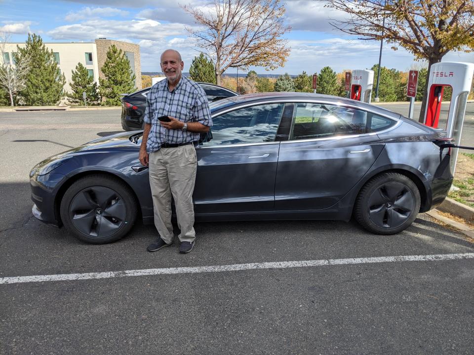 A man stands in front of a Tesla EV charging at a Tesla charger.