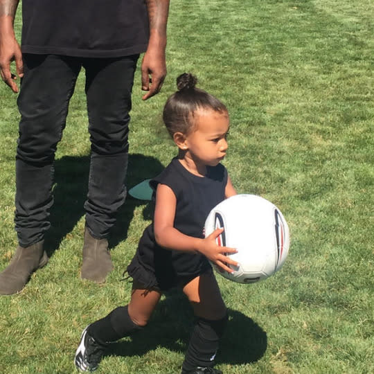 North West playing soccer while Kanye lingers in the background on the field.