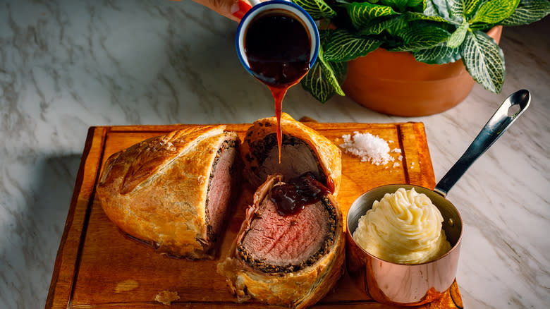 beef wellington on a board with gravy being poured over and a pot of mash