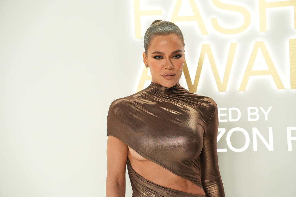 Khloe Kardashian poses on the red carpet in a skin-tight metallic wrap dress that is open over her abs, and a slicked-back bun and dark eyeliner