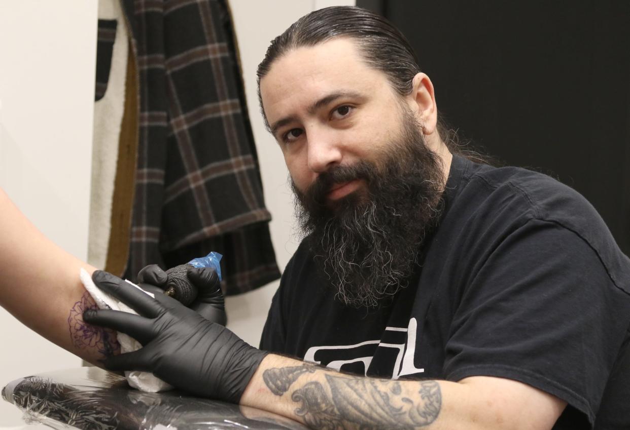 Tattoo artist Dennis Hamilton pauses for a photo as he creates flowers on the arm of Maggie Frazier Saturday afternoon, Jan. 20, 2024, at the Studios on Main Street in Mount Holly.