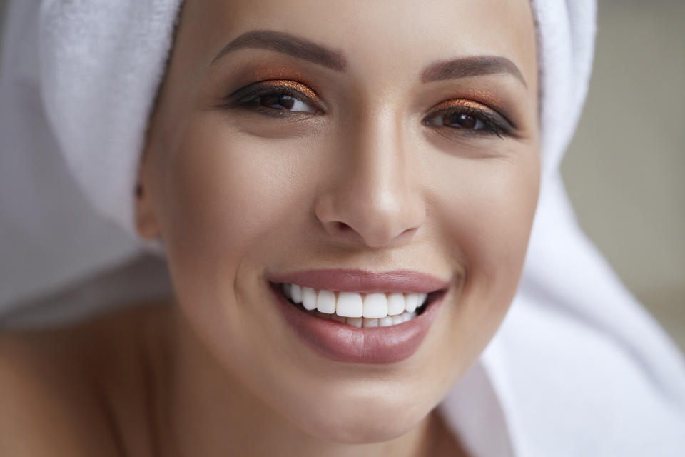 A woman with neatly groomed eyebrows, shimmering eyeshadow, and a radiant smile is wearing a towel wrapped around her head