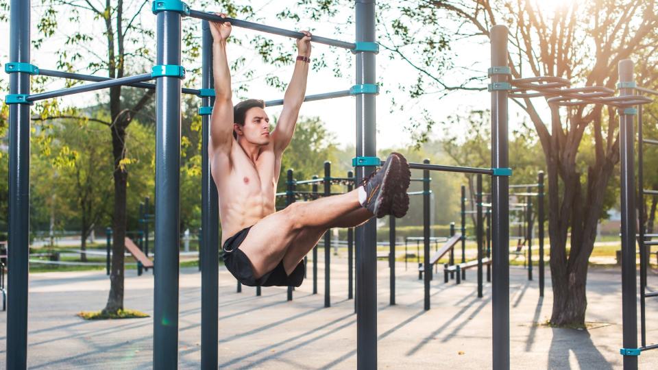 Man performing hanging leg raises in a park with legs extended in front of him, holding on to a bar