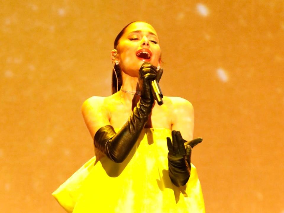 Ariana Grande wearing black opera gloves and an oversized yellow dress performing onstage