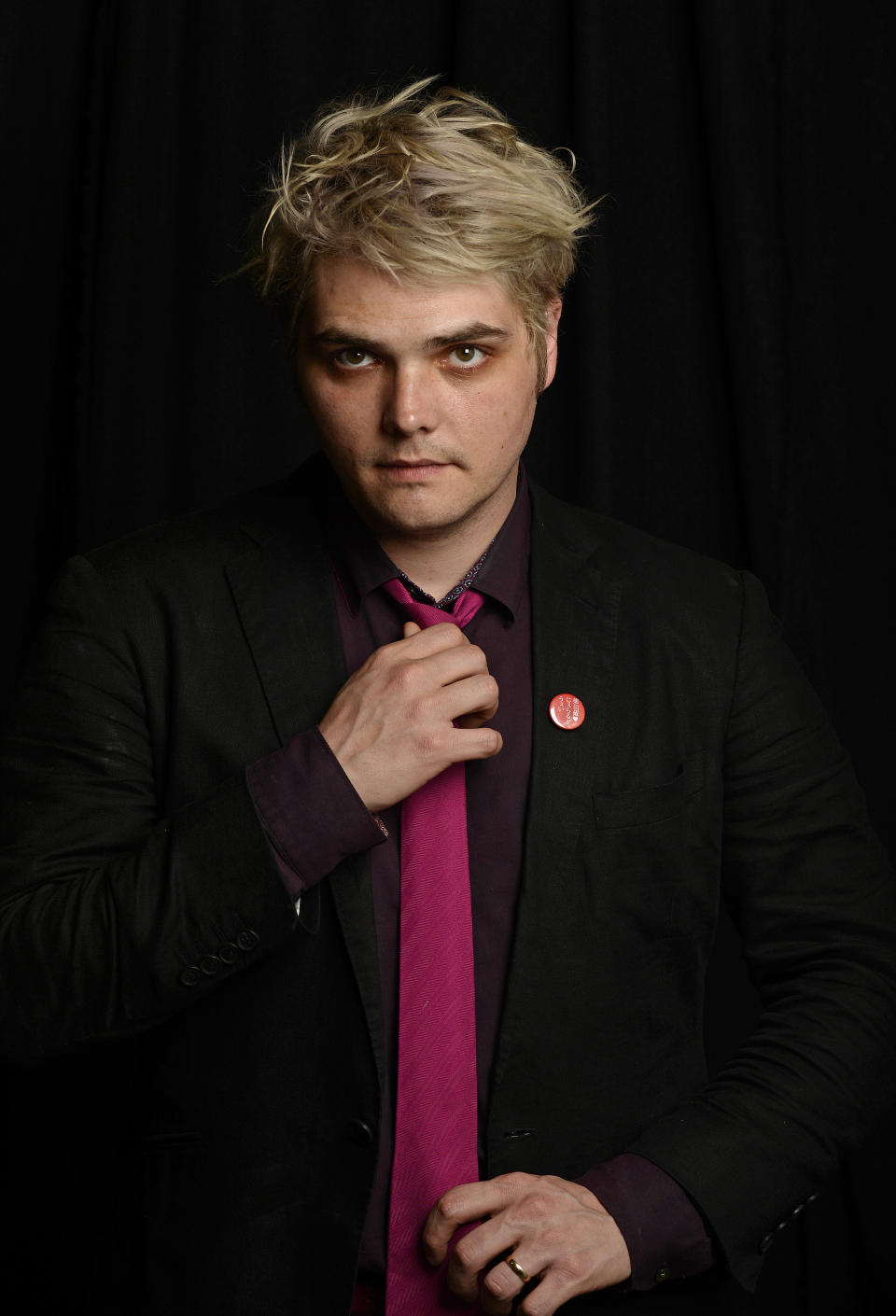 Gerard Way is adjusting his tie while wearing a black suit, purple shirt, and pink tie, with a pin on his lapel. He stands against a dark background