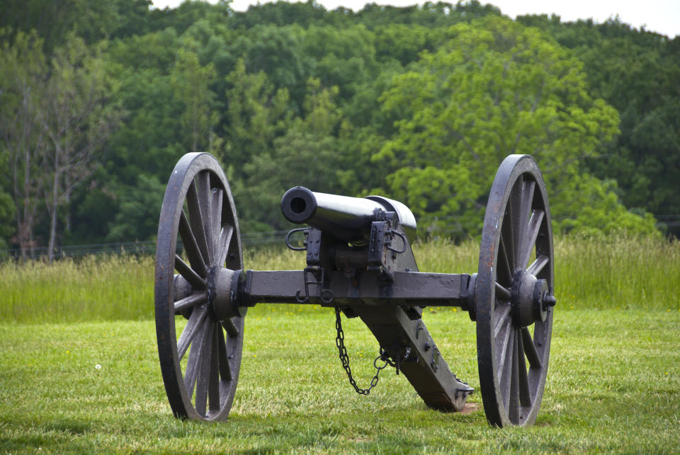 A historic cannon with large wooden wheels is set on a grassy field with dense trees in the background