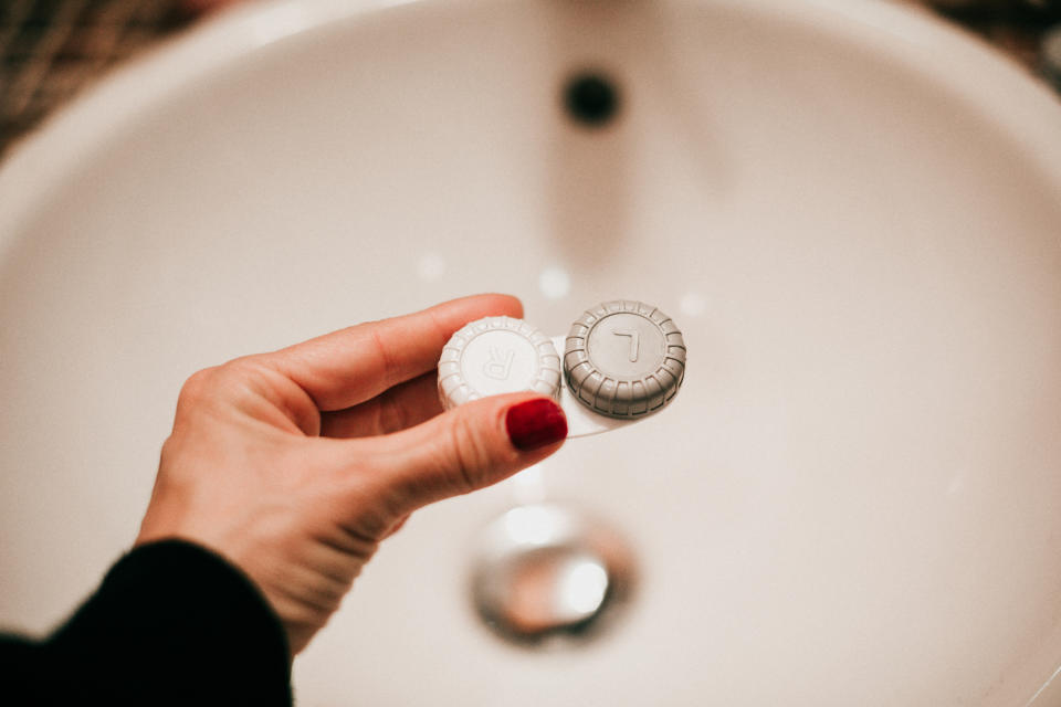 Una mano con le unghie dipinte di rosso tiene una custodia per lenti a contatto sopra il lavandino del bagno