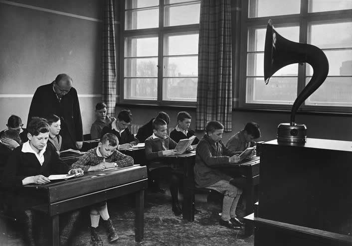 A historical photo of a classroom with students reading books and a teacher supervising, with a phonograph on the desk