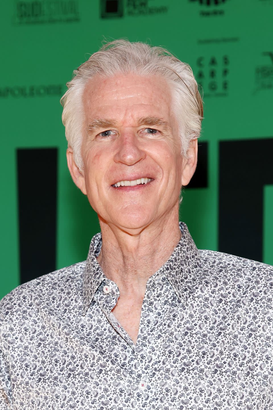 matthew modine smiles at the camera, he wears a black and white paisley patterned shirt