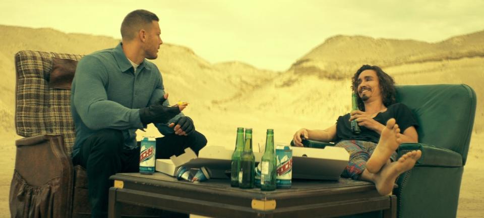 Luther and Klaus sit on couches eating pizza and drinking bear while talking in the middle of a desert