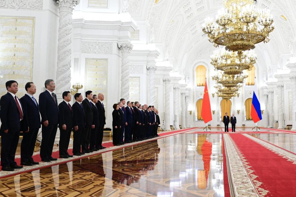 People in dark suits, left, stand in line, awaiting two men standing before a red carpet and under a row of chandeliers