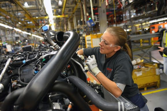 A worker is shown using a tool on an assembled engine, near a production line in a Ford factory.