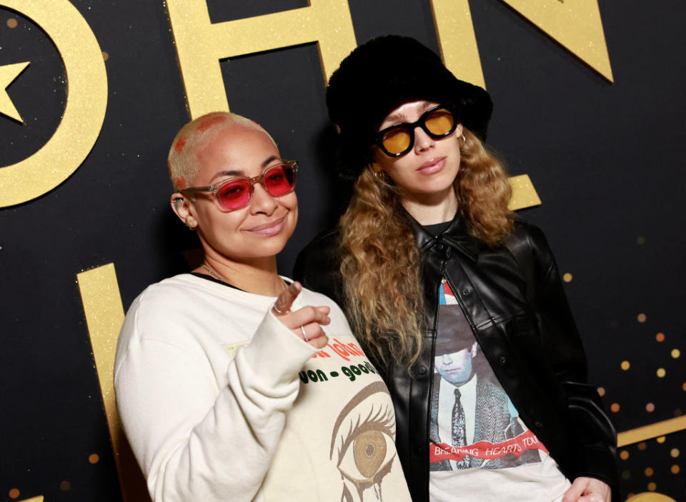 Two celebrities pose together; one in a graphic sweatshirt and beanie, the other in a leather jacket and graphic tee