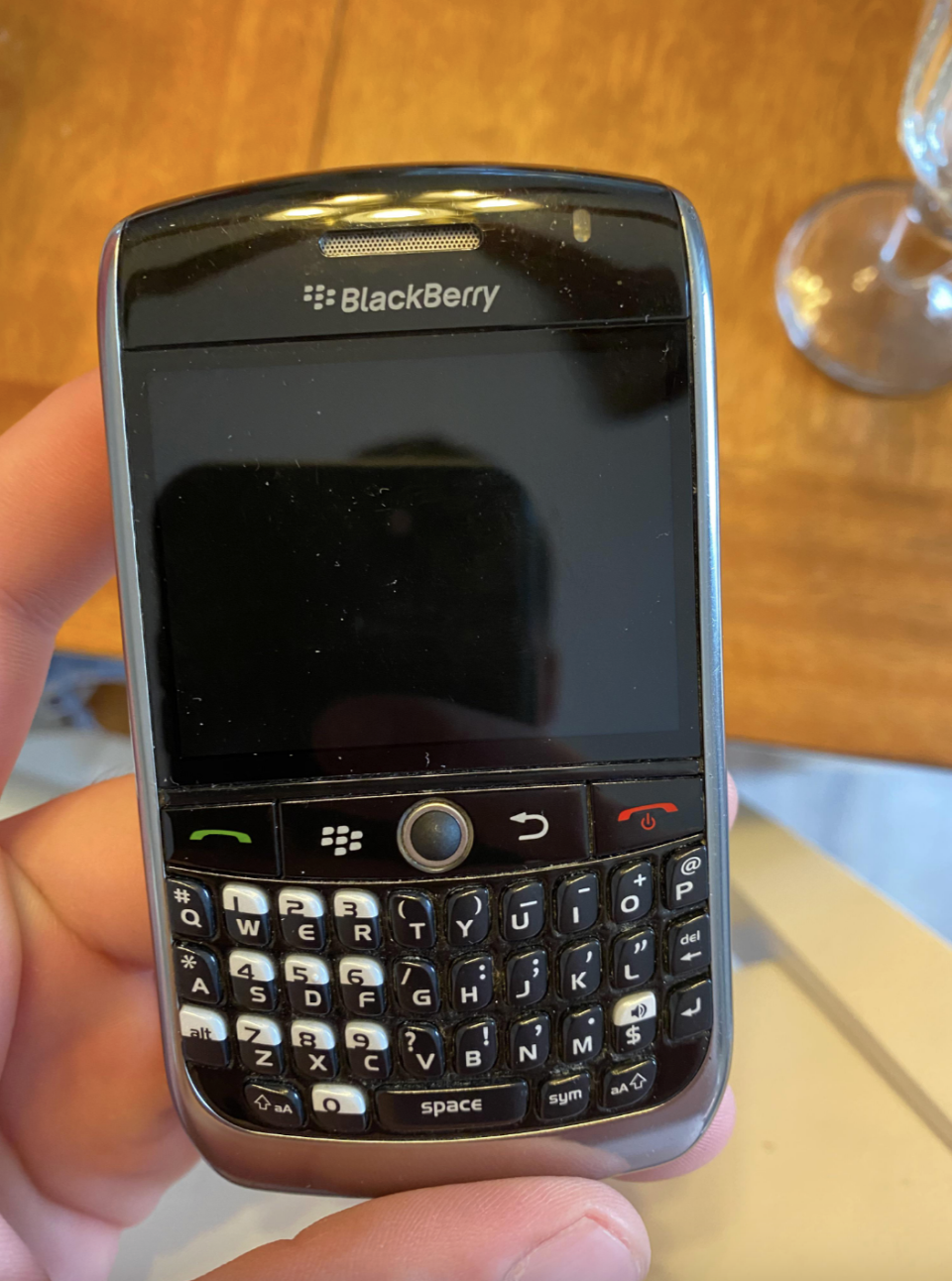 A hand holds a classic BlackBerry smartphone with a physical QWERTY keyboard and a small screen