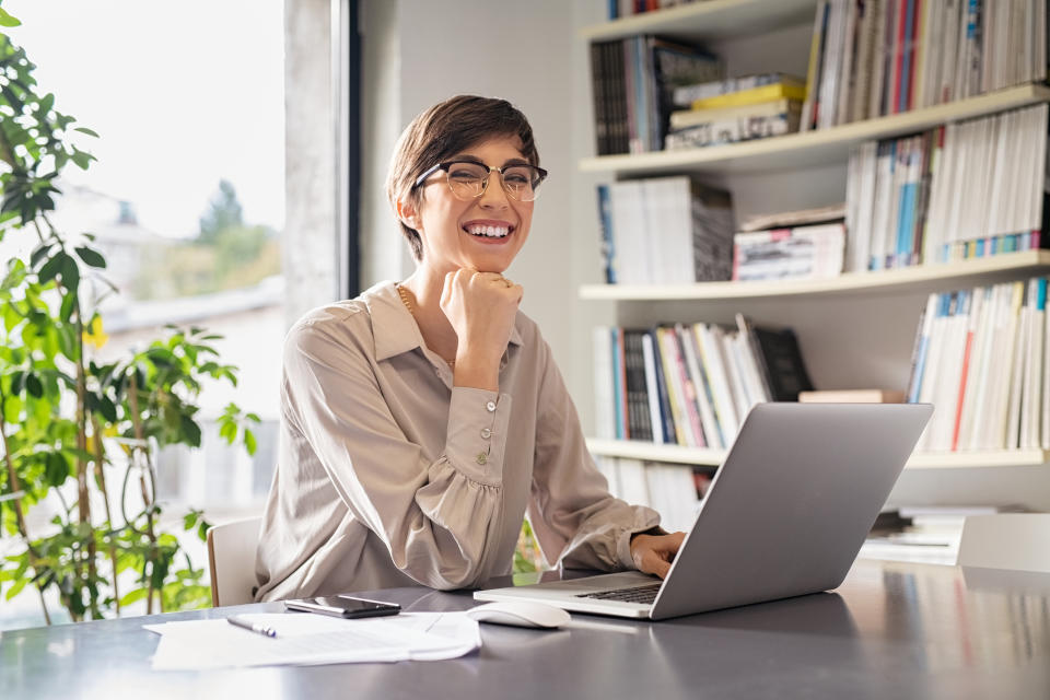 Successful young business woman sitting in creative office and looking at camera. Portrait of happy entrepreneur with hand on chin working on computer. Smiling businesswoman using laptop while working from home.