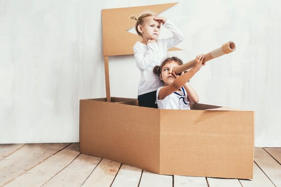 Two little girls playing inside a cardboard box boat, with one seated while looking through a cardboard tube.