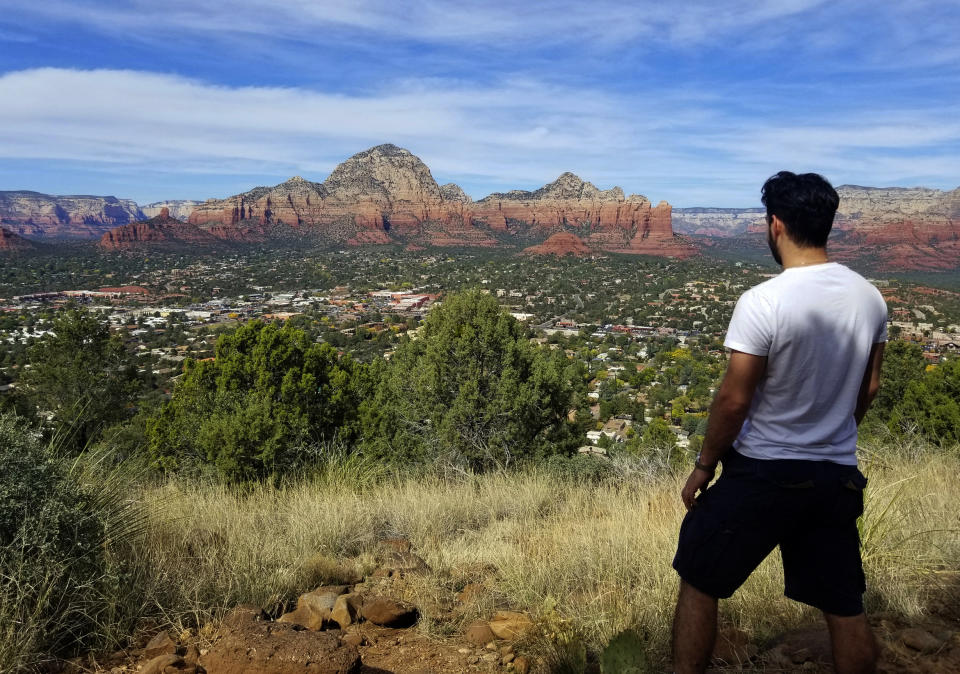 In this 2018 photo provided by Regina de Heer, Joseph Gedeon looks over the town of Sedona, Ariz., seen from the Airport Mesa Loop Trail. From left to right are Chimney Rock, Thunder Mountain, Sugar Loaf and Coffee Pot Rock. The sleepy Arizona town of Sedona has long been a refuge for hikers, romantics and soul searchers. There’s picturesque beauty in its earth-toned buildings and also in the glowing red rocks that surround town. (Regina de Heer via AP)