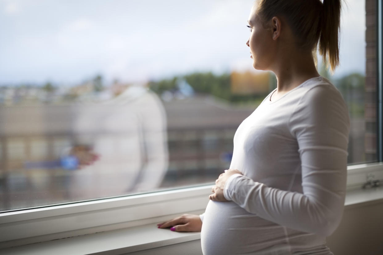 Pregnant woman with prenatal depression looks out the window home. Holding a hand on the tummy. Mental health and pregnancy.