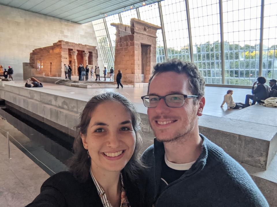 Photo of India and Sam at the Metropolitan Museum of Art in New York City. India has her hair tied back and wears a white shirt with black stripes and flowers, underneath a black blazer. Sam wears a green wool tip and black and white t-shirt. Behind them is an exhibition of Egyptian pyramid-type buildings. Behind that is a wide window panel that stretches from one side of the building to the other.