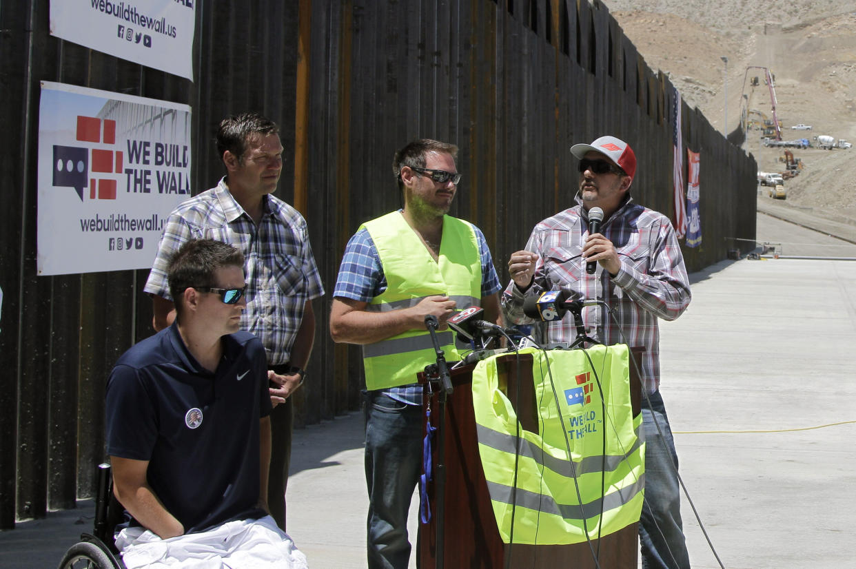 Leaders of We Build the Wall Inc. congregate around a podium with news microphones in front of a wall near a desert landscape.