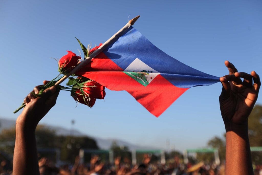 A woman holds a Haitian flag and roses as people pray together during a church service that is set up outdoors near an encampment after numerous churches were destroyed during the massive earthquake on January 24, 2010 in Port-au-Prince, Haiti. (Photo by Joe Raedle/Getty Images)