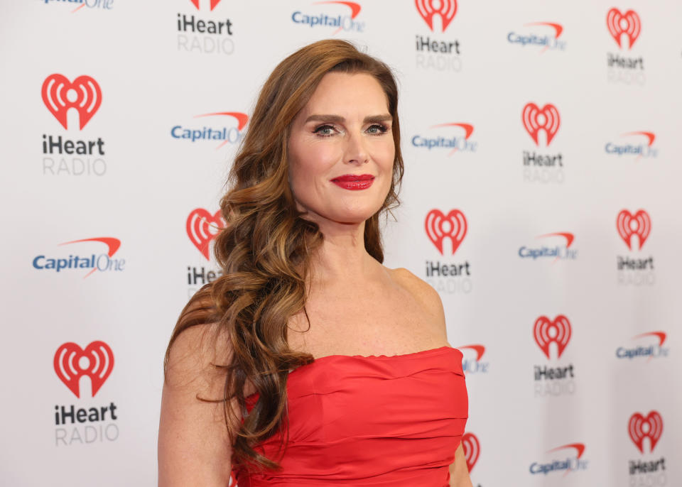 Head and shoulder shot of Brooke smiling, her long medium brown hair worn wavy over one shoulder. She wears a red strapless top and matching red lipstick.