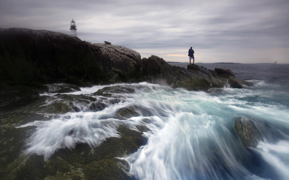 FILE - This July 25, 2011 file photo shows the surf crashing on the rocks as a man fishes for striped bass below Portland Head Light at Fort Williams Park in Cape Elizabeth, Maine. This lighthouse in Cape Elizabeth, complete with a museum and a gift shop, has been called one of the most-photographed lighthouses in the country. Commissioned by George Washington, the light went into service in 1791. It became automated in 1989 and is now owned by the town of Cape Elizabeth. The lighthouse is the premier attraction at Fort Williams Park, a six-mile drive from Portland. (AP Photo/Robert F. Bukaty, file)