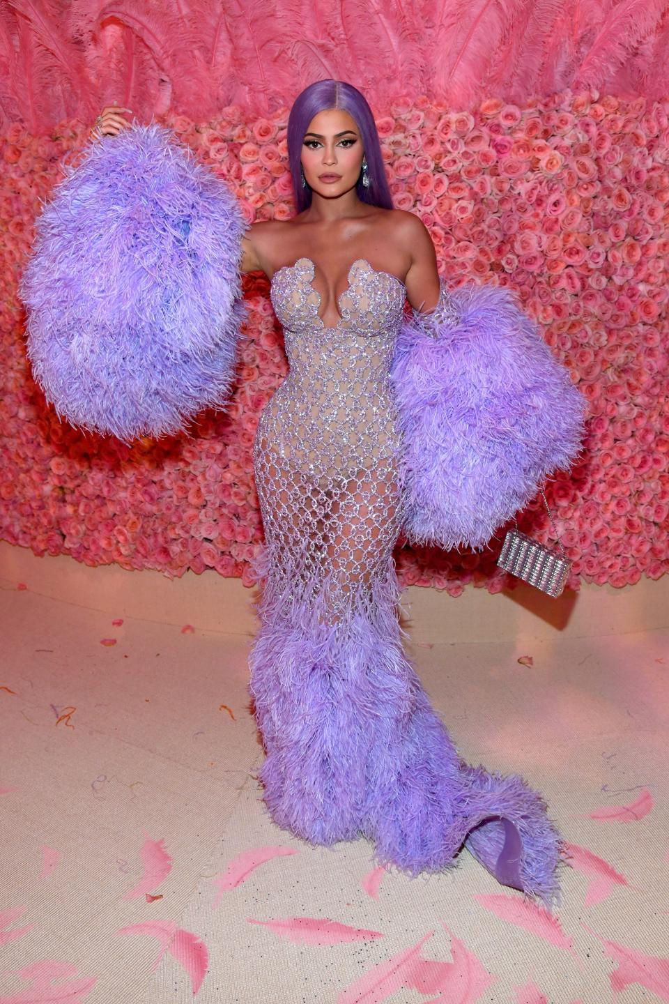 Kylie Jenner poses in front of a wall of pink roses wearing a shimmering dress with a purple feathered skirt, giant purple pom poms, and purple hair.