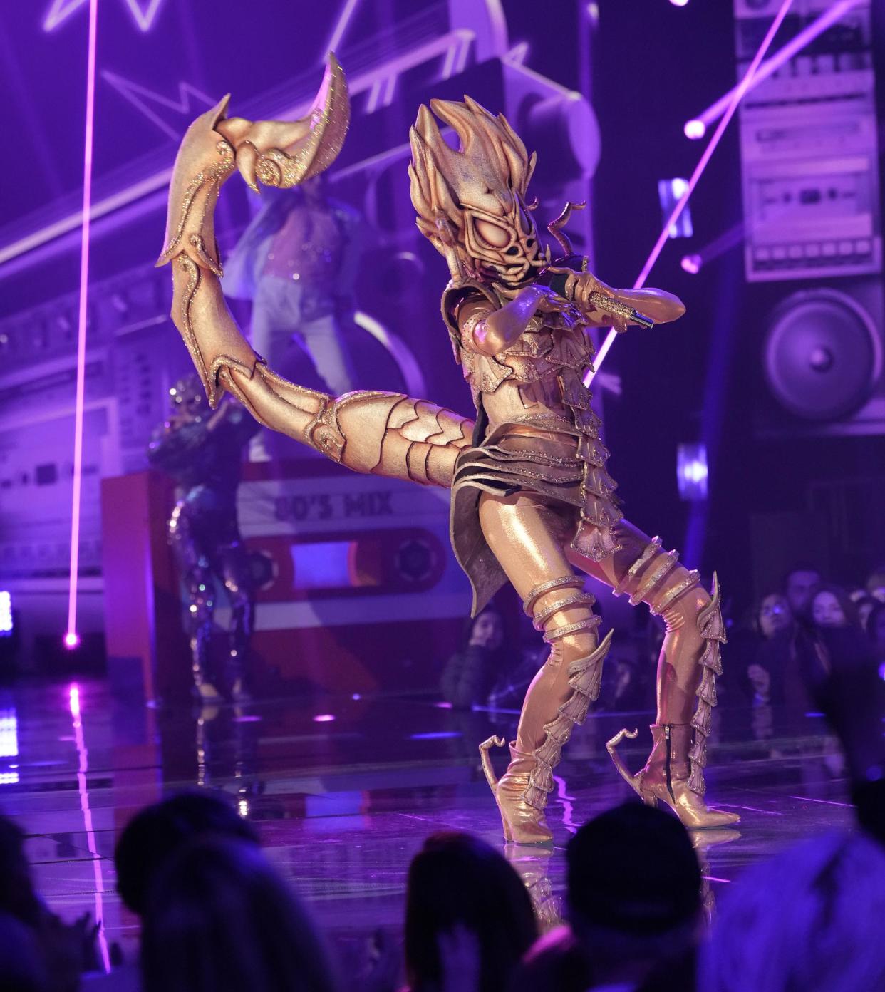 A person in a gold sparkly scorpion costume stands on stage holding a mic against a purple backdrop. (Michael Becker / FOX)