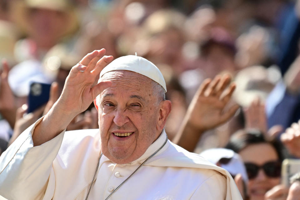 A smiling Pope Francis waves to a crowd, wearing his traditional white papal vestments and skullcap, during a public appearance