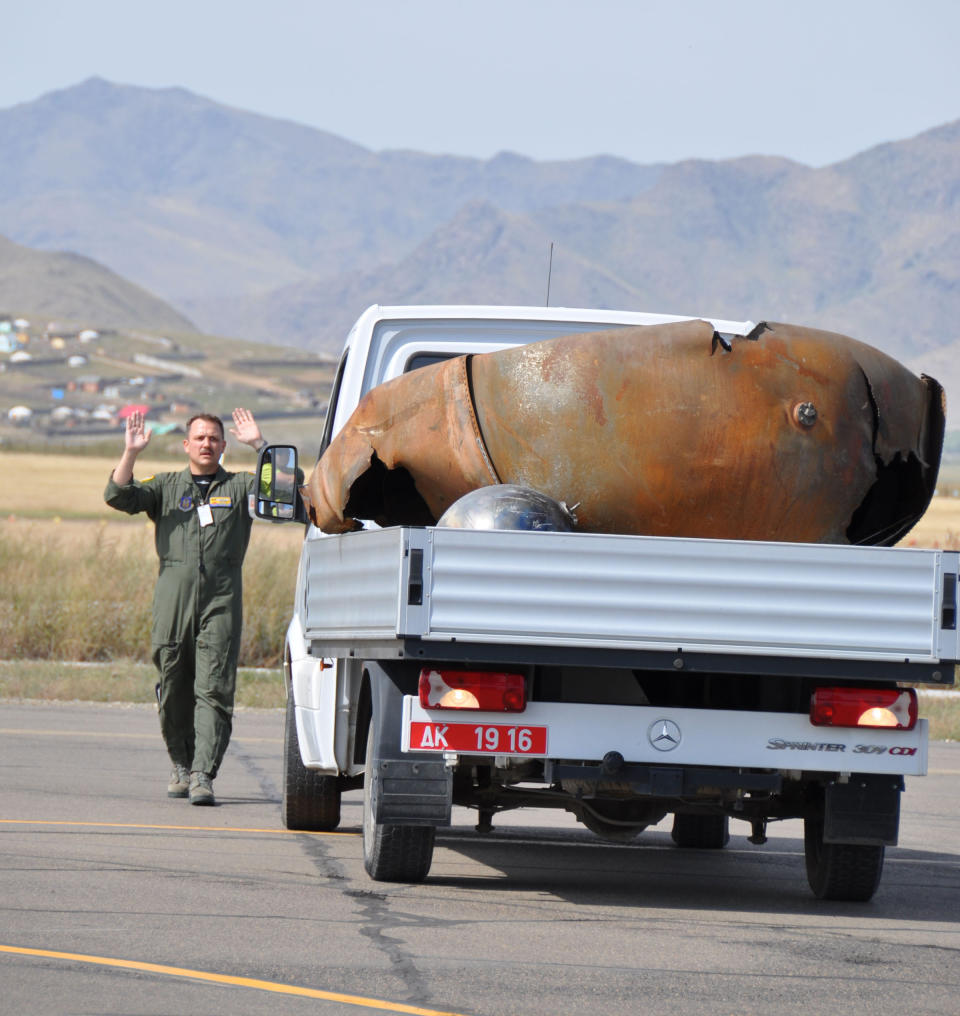 A man in a green jumpsuit raises his arms next to a white utility vehicle containing a dilapidated metal shard in its bed.