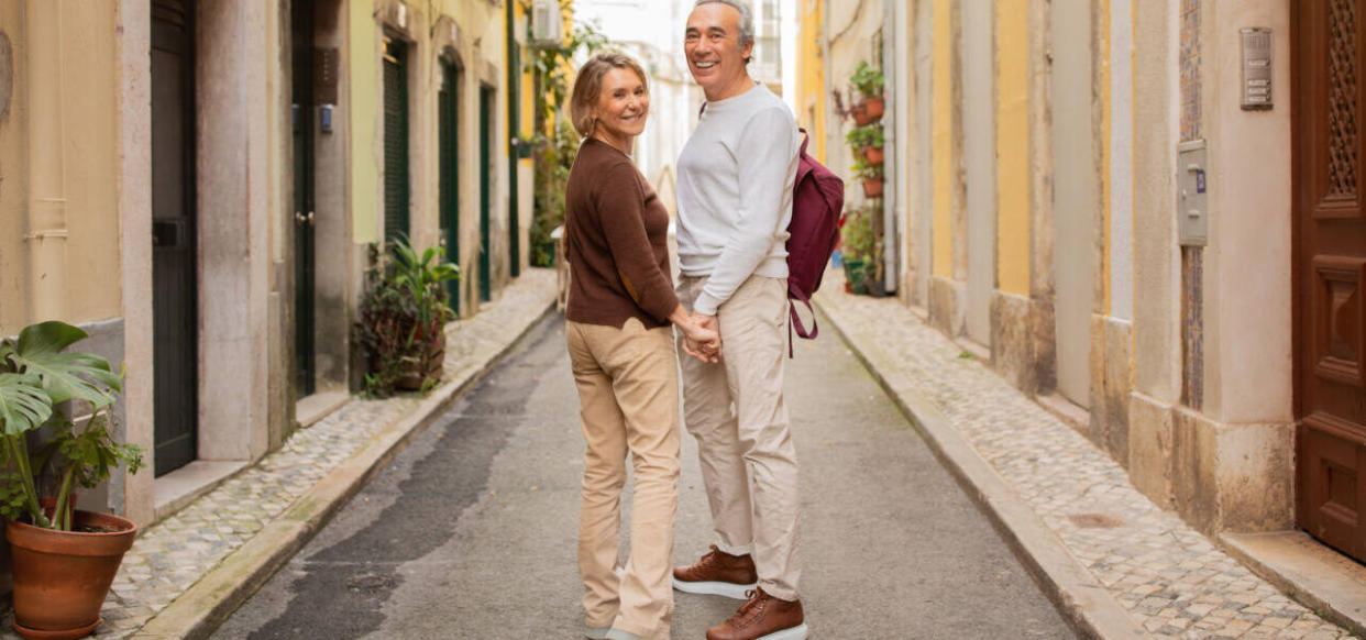 Vacation In Europe. Happy Mature Couple Traveling Standing With Backpack And Holding Hands Smiling To Camera Posing Outside On Lisbon Street. Travel And Tourism, Sightseeing Tour Offer Concept